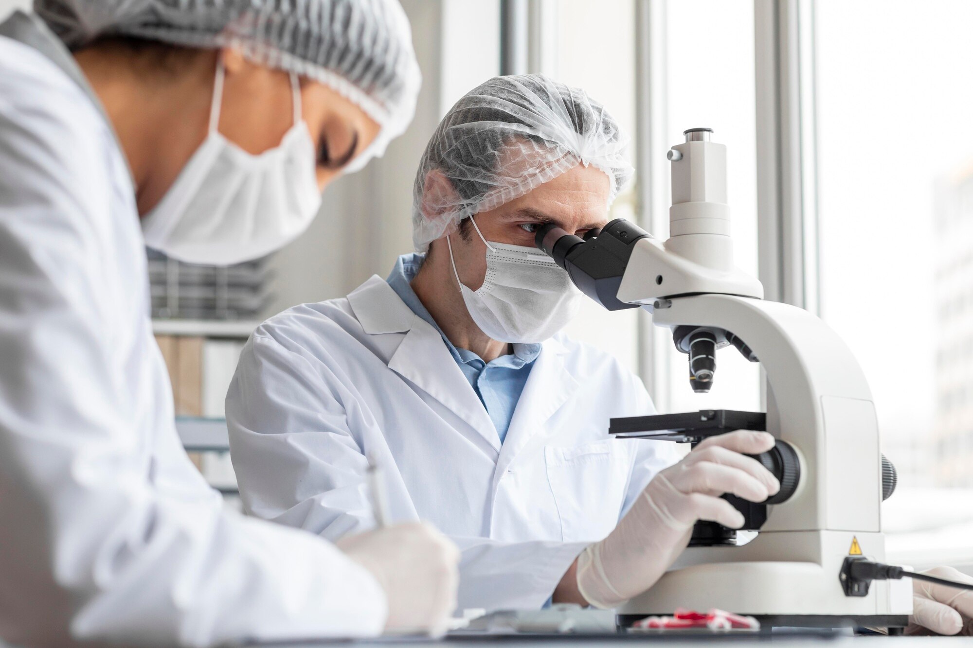 A male scientists wearing a cap and mask looking through a microscope in a lab